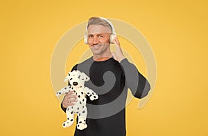 Happy infantile middle-aged man play with toy dog listening to music yellow background, headphones