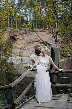 Happy interracial couple newlyweds stands on wooden bridge and embraces against background of lake, forest and canyon