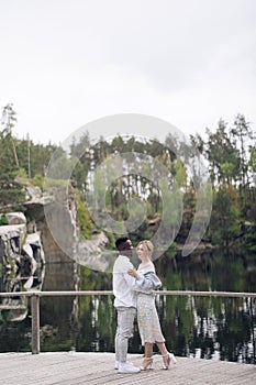 Happy interracial couple stands on wooden bridge and embraces against background of lake, rocks and forest
