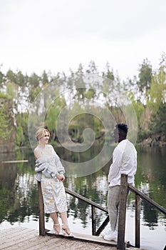 Happy interracial couple stands on wooden bridge, looks each other and smiles against background of lake and forest