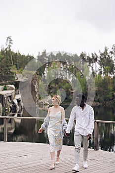 Happy interracial couple walks on wooden bridge and holds each other hands against background of lake, rocks and forest