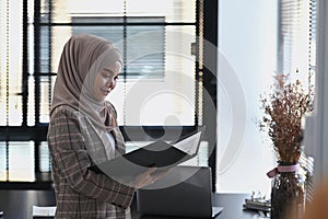 A happy islamic girl in hijab is inspecting and approving documents in folder and standing beside windowpanes.