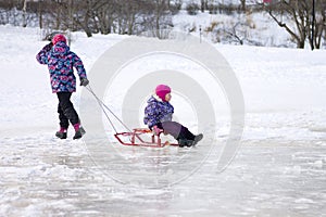 Happy ittle girl pulling her young sister on a sled on the ice in snowy winter park