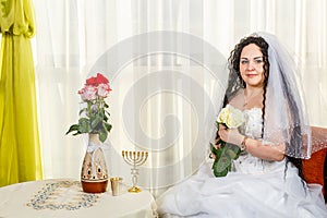 A happy Jewish bride with a bouquet of white roses sits in the synagogue before the Huppa ceremony at a table with