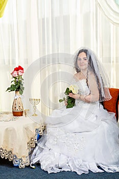 A happy Jewish bride with her face covered with a veil with a bouquet of white roses sits in a synagogue before