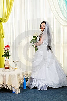 A happy Jewish bride stands in the hall before the chuppa ceremony at a table with flowers with a bouquet of white roses