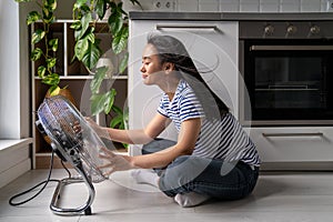 Happy joyful Asian girl sitting in front of electric fan, refreshing during hot summer days at home