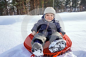 Happy joyful beautiful child boy rides from the mountain on a red tubing in winter