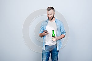 Happy joyful cheerful bachelor is drinking beer and using smartphone for typing sms, isolated on grey background