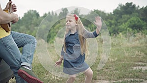 Happy joyful little girl dancing having fun as unrecognizable man playing ukulele sitting in car trunk on summer meadow