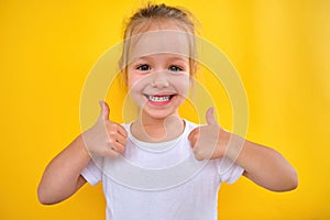 Happy joyful little preschool girl smiling showing thumbs up yellow background