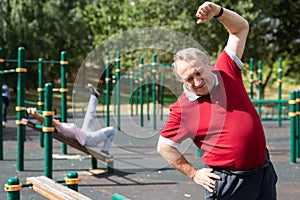 Happy joyful retired sportsman in sportswear doing exercises with arm above head, exercising outdoors in city park