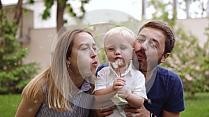 Happy, joyful small child in the middle. Parents close to the boy from both sides. The are playfully blowing dandelion