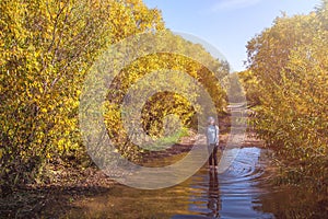 A happy joyful smiling contented child is standing in a puddle on a dirt road in a forest in autumn in sunny weather.