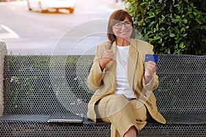 Happy joyful smiling freelancer adult woman 50-60 s wearing stylish suit is holding credit card she excited raised hands