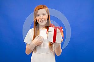 Happy joyful teenager girl with red hair does not believe happiness holding a gift box on a blue studio background