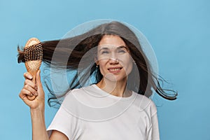 happy, joyful woman combing her long, beautiful, well-groomed hair with a wooden comb in a white t-shirt on a light blue