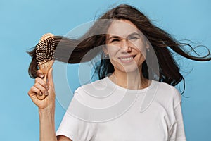 happy, joyful woman combing her long, beautiful, well-groomed hair with a wooden comb in a white t-shirt on a light blue