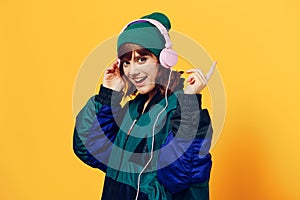 a happy, joyful woman in a green jacket and hat enjoys music standing in pink headphones on a yellow background raising