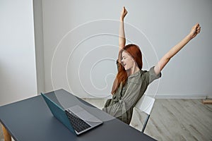 happy, joyful woman sitting at a table with a laptop rejoices raising her hands up and looking at the camera
