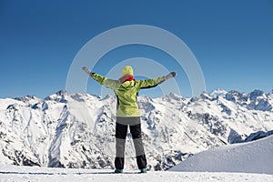 Happy joyful woman in ski suit having fun outdoors in winter, standing on the mountains covered with snow with raised up