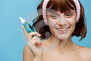 a happy, joyful woman stands on a blue background with a jar of facial serum with a soft pink rim on her head and smiles