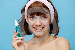 a happy, joyful woman stands on a blue background with a jar of facial serum with a soft pink rim on her head and smiles