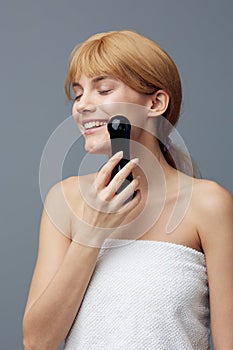 a happy, joyful woman stands on a gray background wrapped in a towel and does a facial massage with a black electric