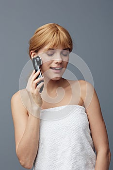 a happy, joyful woman stands on a gray background wrapped in a towel and does a facial massage with a black electric