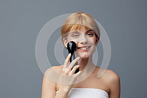 a happy, joyful woman stands on a gray background wrapped in a towel and does a facial massage with a black electric