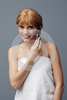 a happy, joyful woman stands on a gray background wrapped in a towel and does a facial massage with a black electric