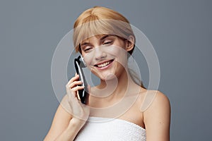 a happy, joyful woman stands on a gray background wrapped in a towel and does a facial massage with a black electric