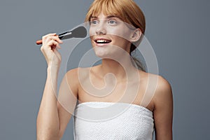 a happy, joyful woman stands on a gray background wrapped in a towel and paints her face with a fluffy makeup brush