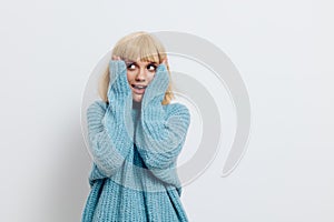 a happy, joyful woman stands on a light background in a blue sweater and looks up in surprise with her hands on her face