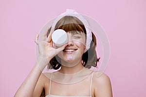 a happy, joyful woman stands on a pink background, with a smeared cream on her cheek and happily smiling holding a white