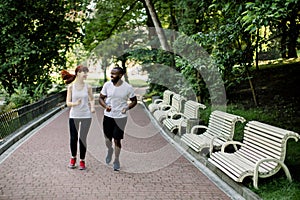 Happy joyful young multiethnic couple, African American man and Caucasian woman, jogging across the city park alley at