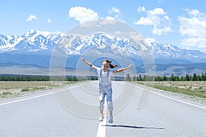 Happy jumping little girl tourist on the beautiful mountain summer empty road. Enjoying traveling. Snow capped mountain