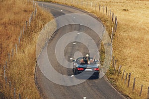 Happy just married couple is driving a convertible car on a country road for their honeymoon, the bride have fun with hands up