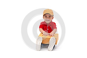 Happy kid Asian boy delivery man in yellow cap and red shirt sitting with parcel post box isolated over white background.