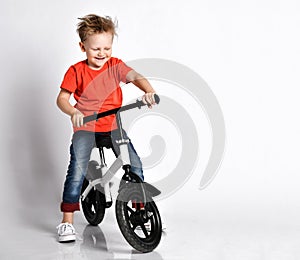 Happy kid boy in orange t-shirt and blue jeans tries to ride his new bicycle without pedals. He tests it turning handlebar