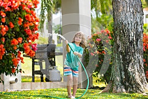 Happy kid boy pours water from a hose. American kids childhood. Child watering flowers in garden. Home gardening.