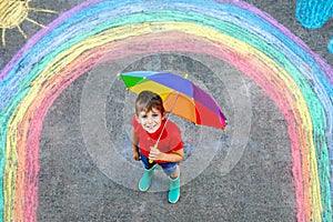 Happy kid boy in rubber boots with rainbow sun and clouds with rain drops painted with colorful chalks on ground or