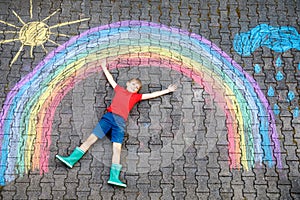 Happy kid boy in rubber boots with rainbow sun and clouds with rain drops painted with colorful chalks on ground or