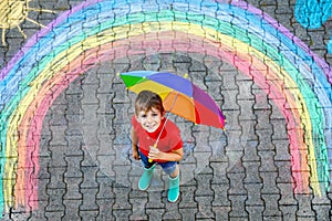 Happy kid boy in rubber boots with rainbow sun and clouds with rain drops painted with colorful chalks on ground or