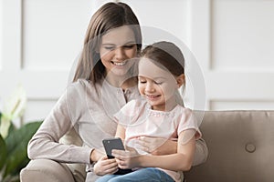 Happy kid daughter and mom enjoy using phone on sofa