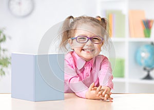 Happy kid girl in eyeglasses reading with standing book on table