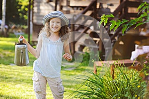 Happy kid girl in hat playing little gardener and helps to water flowers