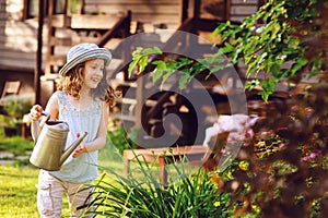 Happy kid girl in hat playing little gardener and helps to water flowers