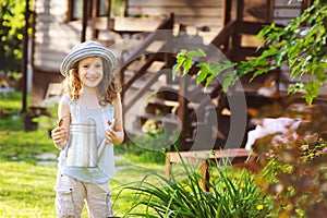 Happy kid girl in hat playing little gardener and helps to water flowers