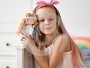 Happy kid girl in home clothing and headband with bow plays takes picture is posing with her favorite doll at home